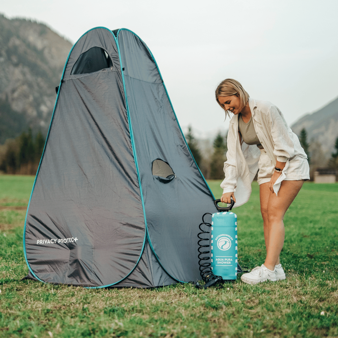Eine junge Frau in kurzen Hosen steht mit einer Campingdusche neben einem dunkelgrauen Duschzelt