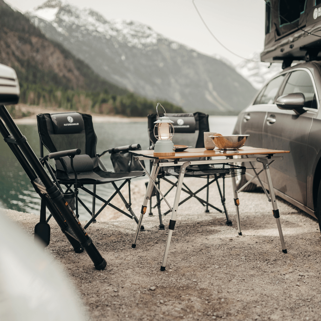 Nahaufnahme eines Campingplatzes mit einem Tisch, Stühlen und einer Lampe. Im Hintergrund befindet sich ein Gebirgszug.