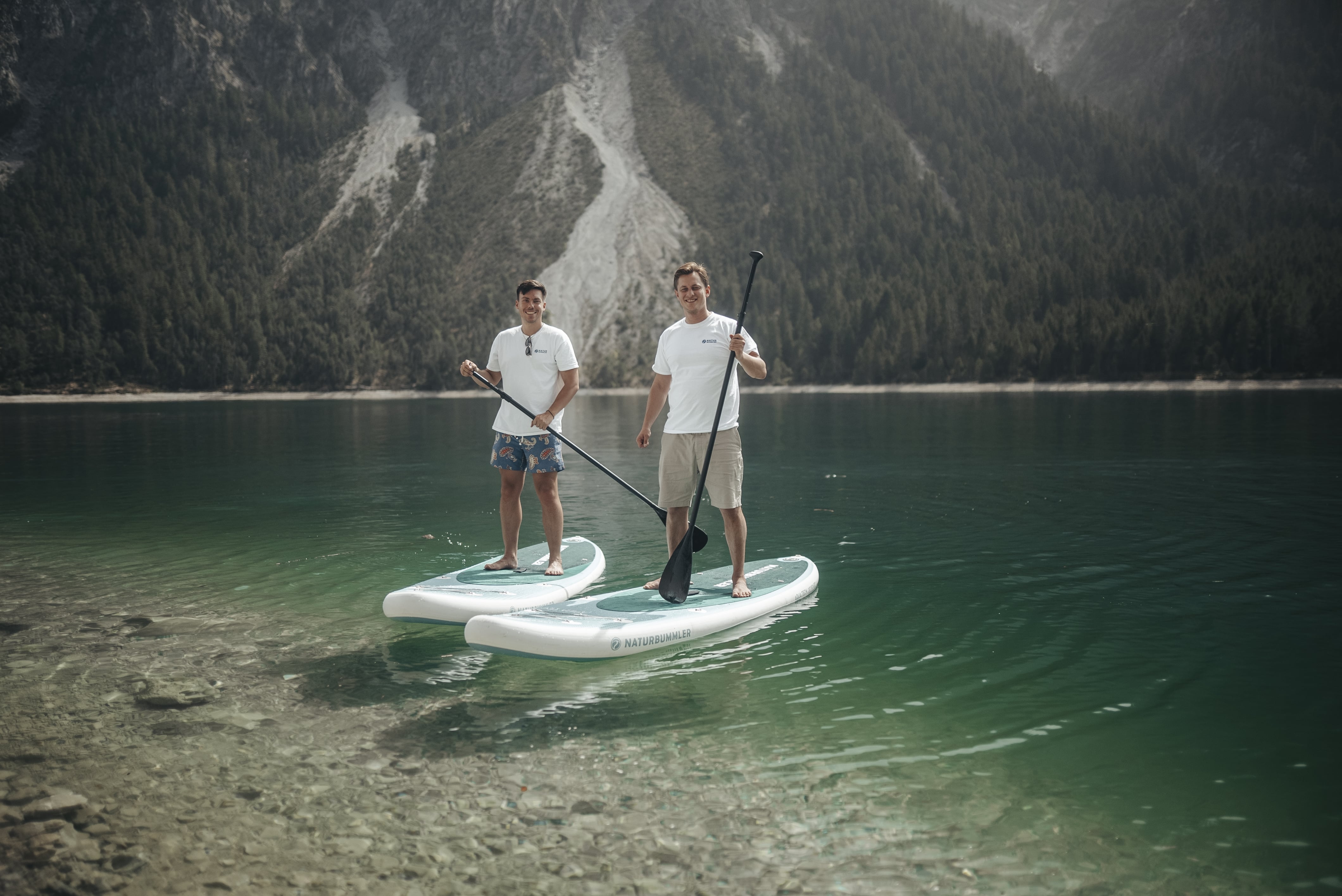 Zwei Männer mit T-Shirts und kurzer Hose stehen auf SUPs auf einem See.
