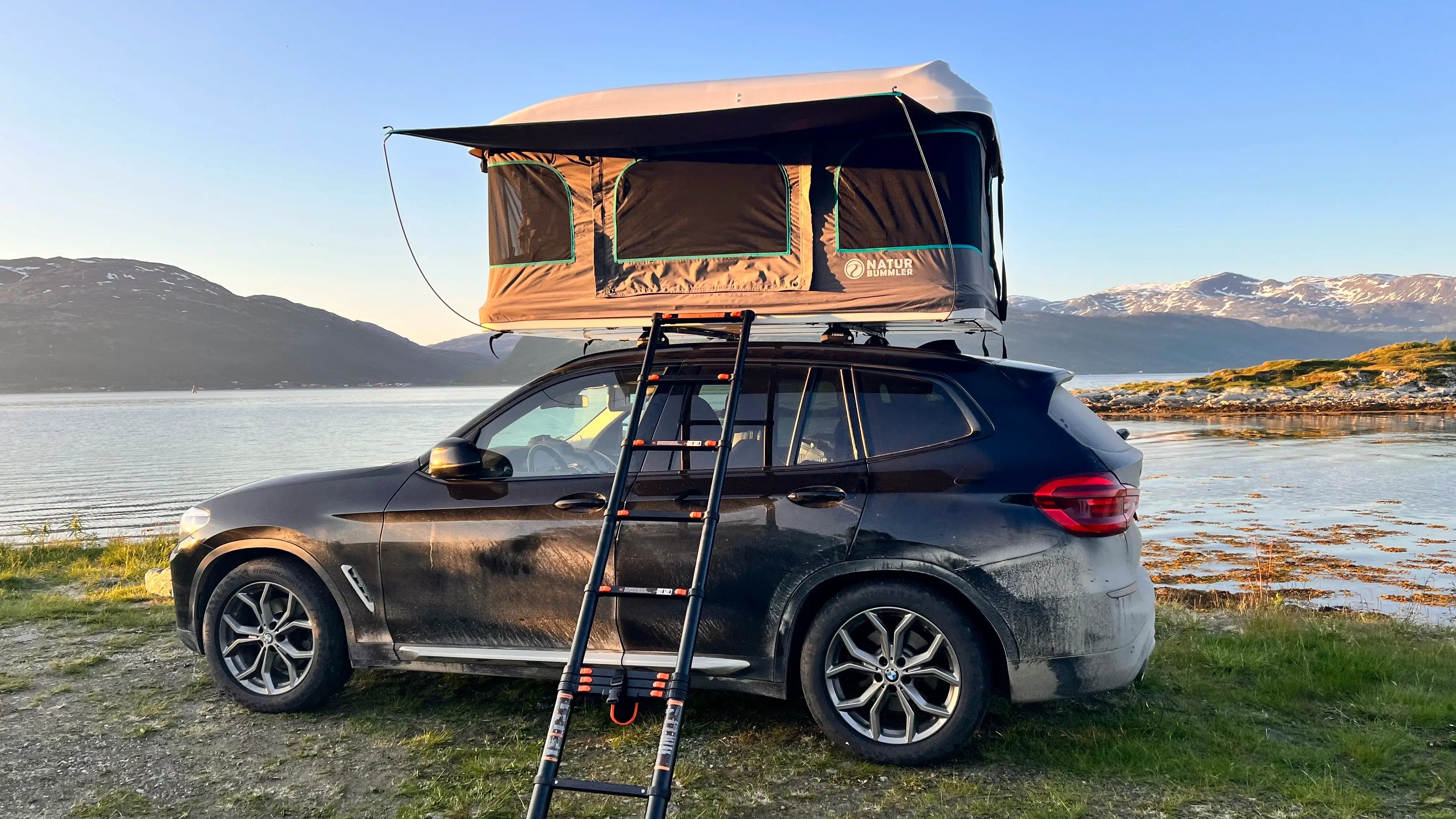 Seitliche Aufnahme eines schwarzen BMWs mit einem aufgestellten Dachzelt vor einem See in den Bergen.